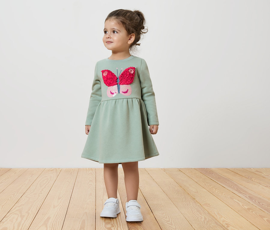 Dívka v zelených šatech s motýlem stojí na dřevěné podlaze.