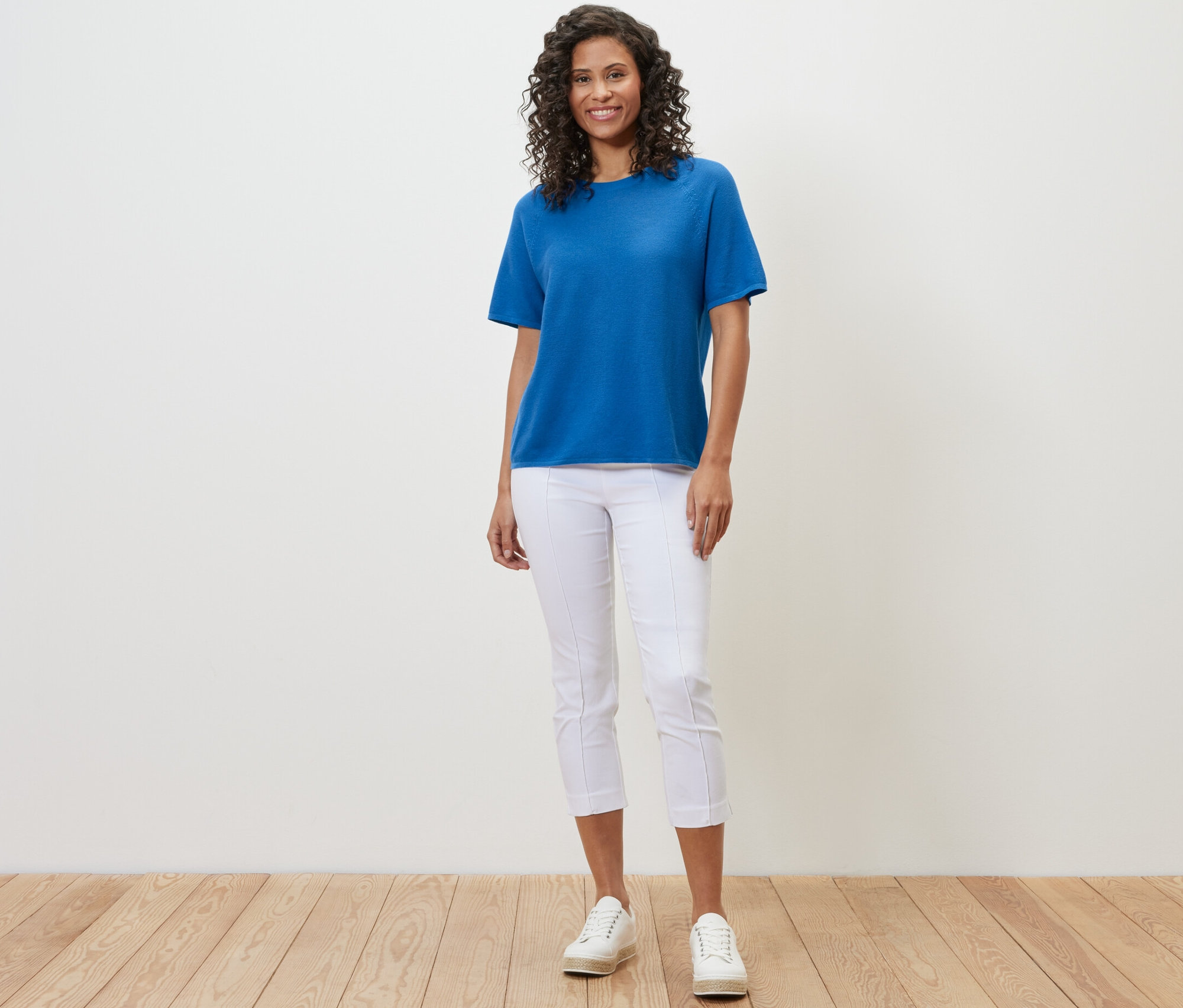 Žena má na sobě kobaltově modré tričko z jemného úpletu, bílé kalhoty a bílé tenisky.