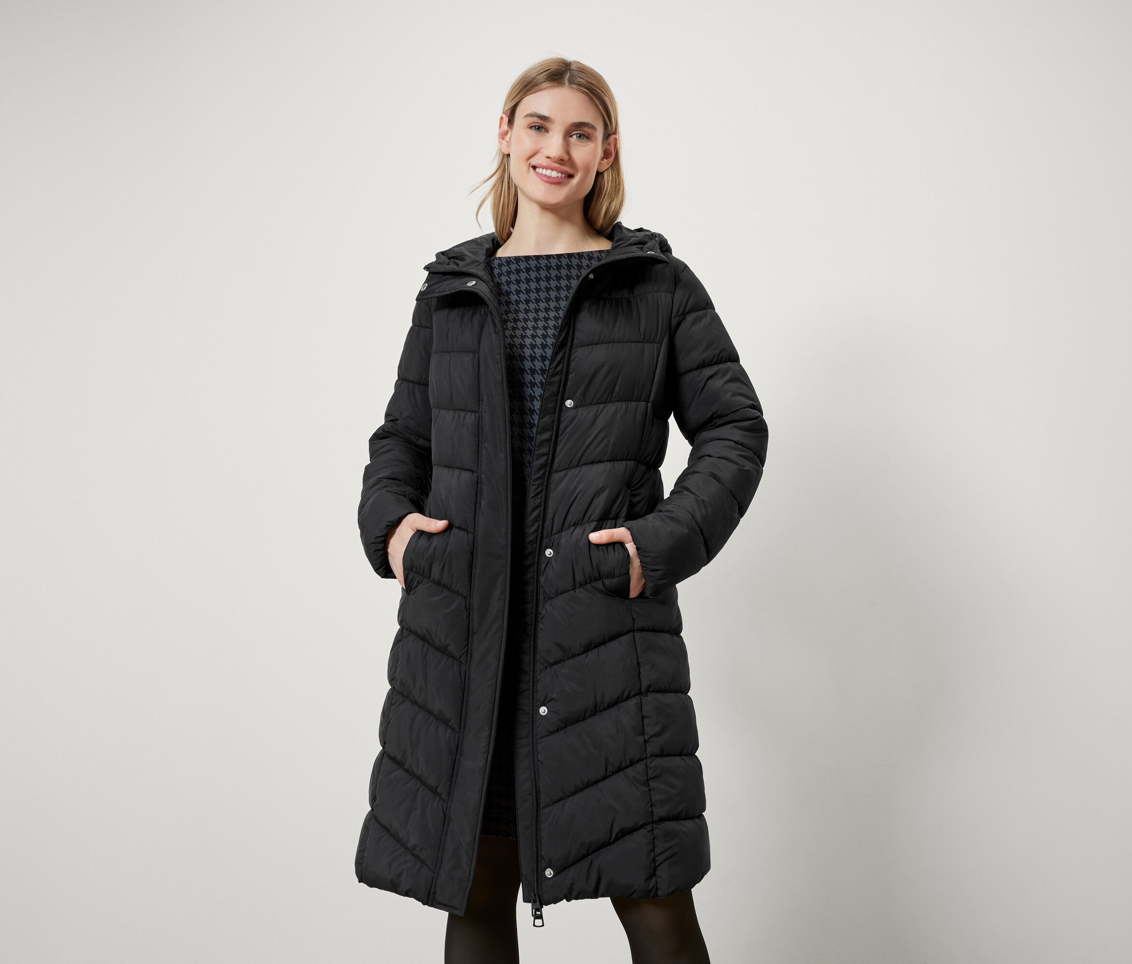 Žena s blond vlasy stojí a má na sobě dlouhou černou prošívanou bundu s kapucí.