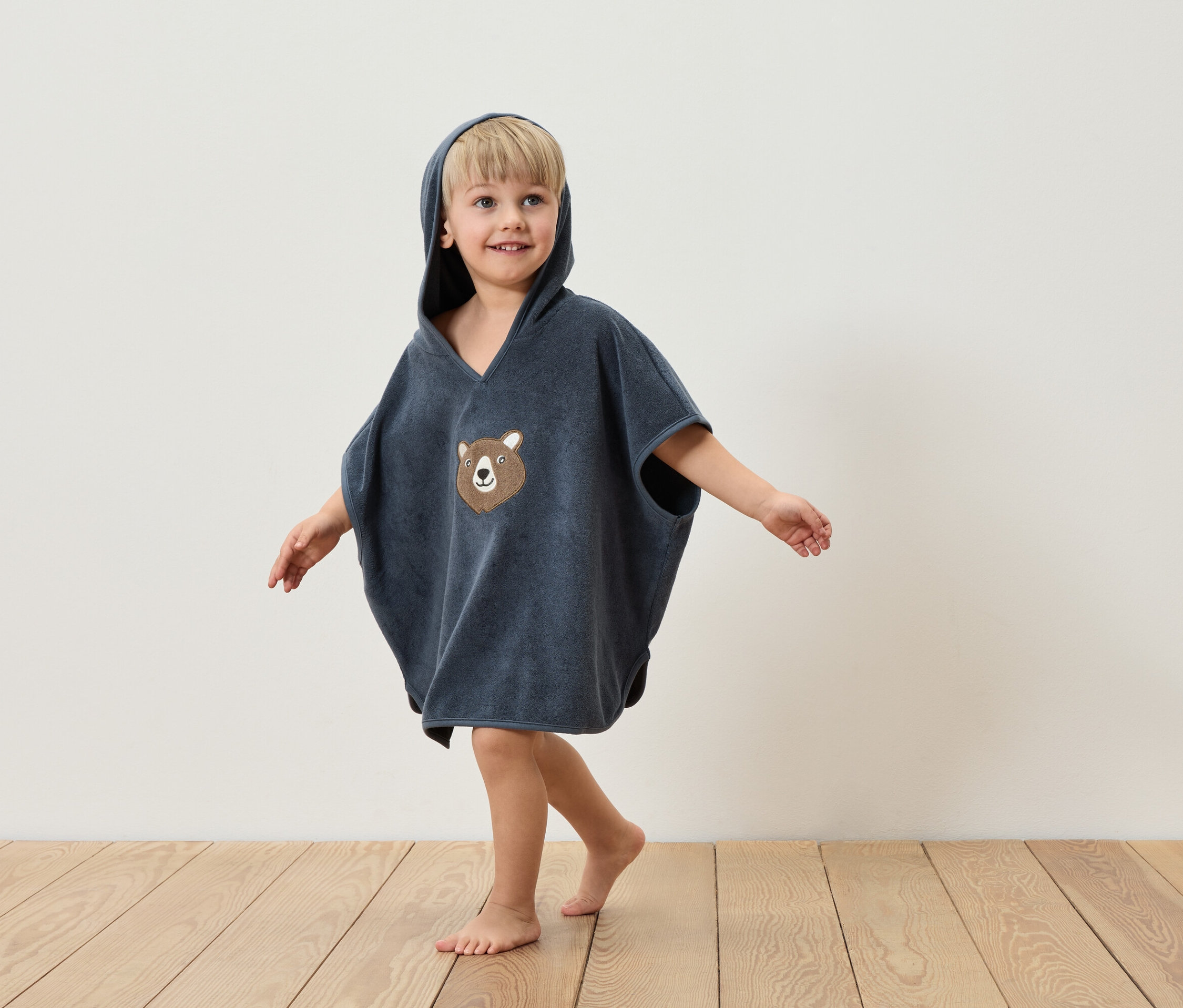 Chlapec v modrém ponču s kapucí a medvědí aplikací stojí na dřevěné podlaze před bílou zdí.