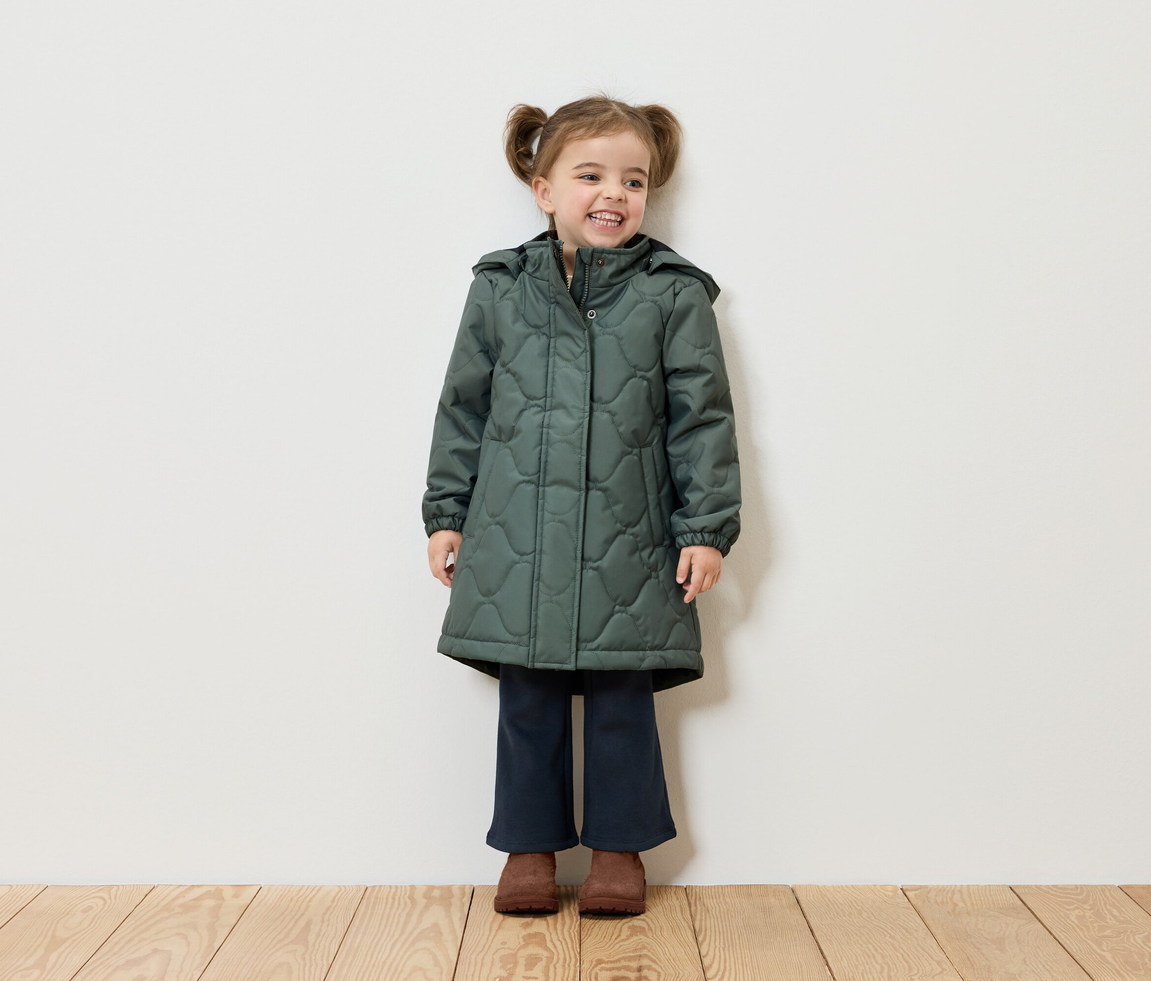 Usmívající se dívka s culíky stojí před bílou zdí a má na sobě zelený kabát, tmavě modré kalhoty a hnědé boty.