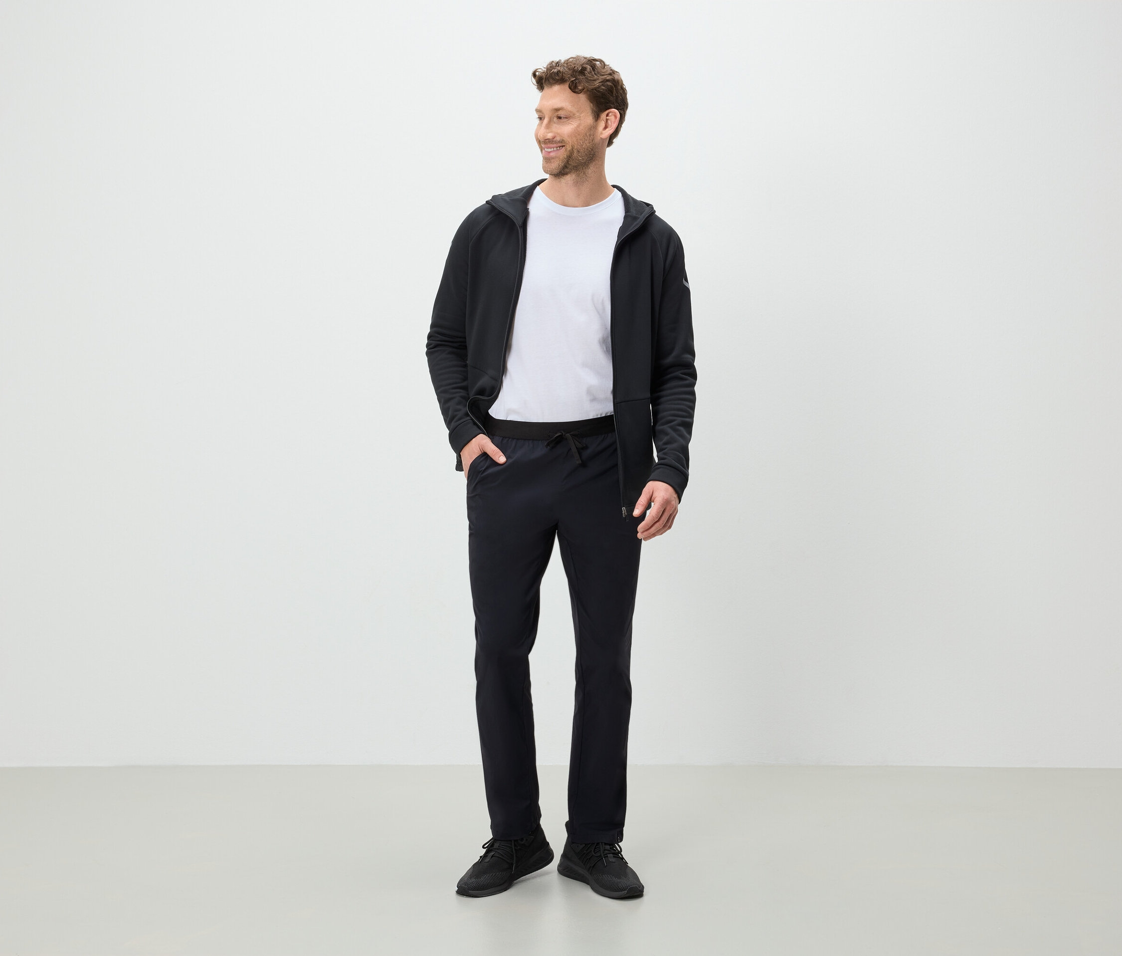 Muž stojí a dívá se stranou, má na sobě černé kalhoty, bílé tričko a černou mikinu s kapucí.