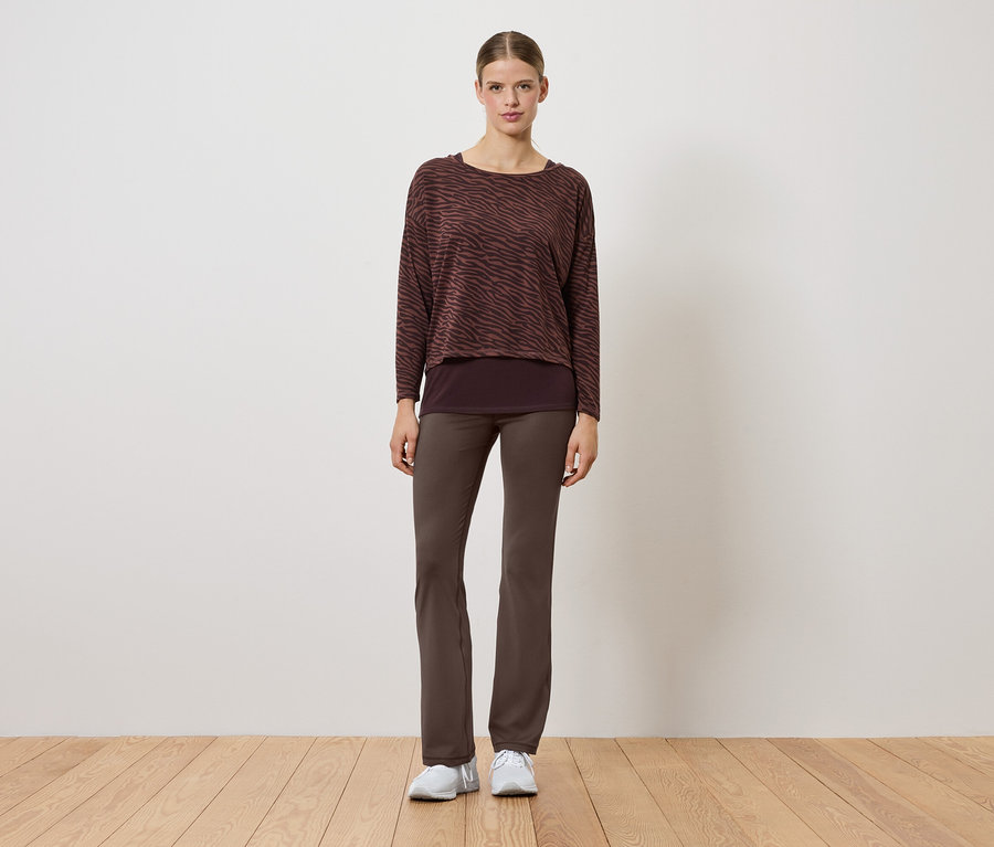 Žena v hnědém svetru a kalhotách stojí na dřevěné podlaze.