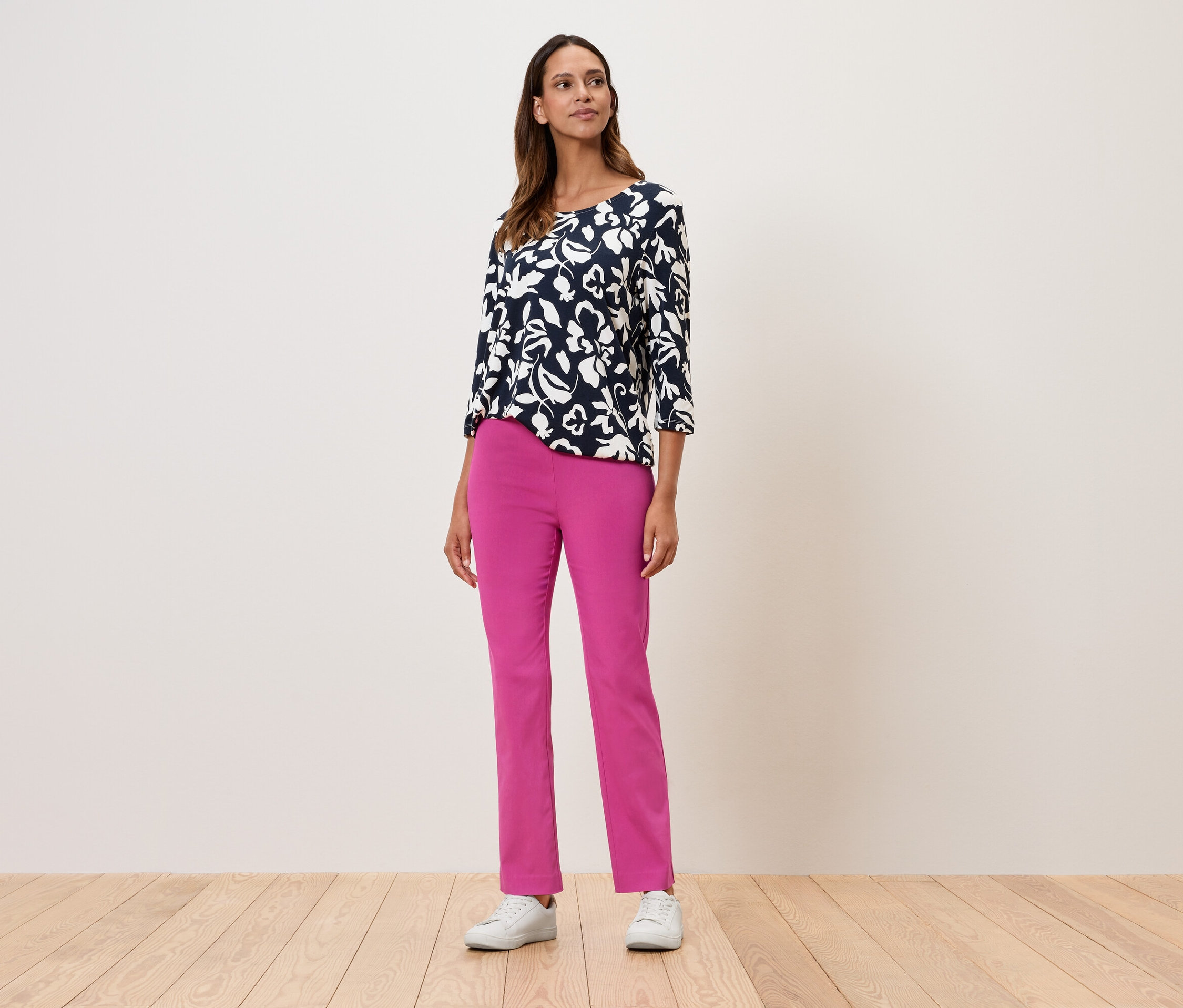 Žena stojí a má na sobě květinový top s tříčtvrtečními rukávy, růžové kalhoty a bílé tenisky.