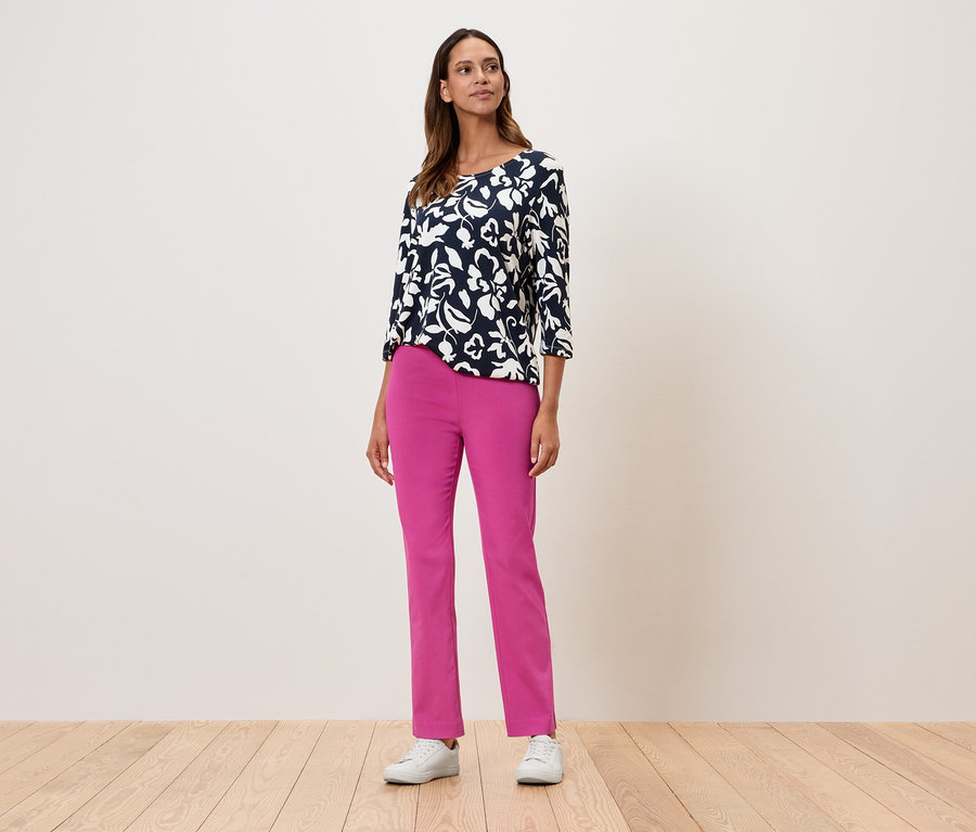 Žena stojí a má na sobě květinový top s tříčtvrtečními rukávy, růžové kalhoty a bílé tenisky.