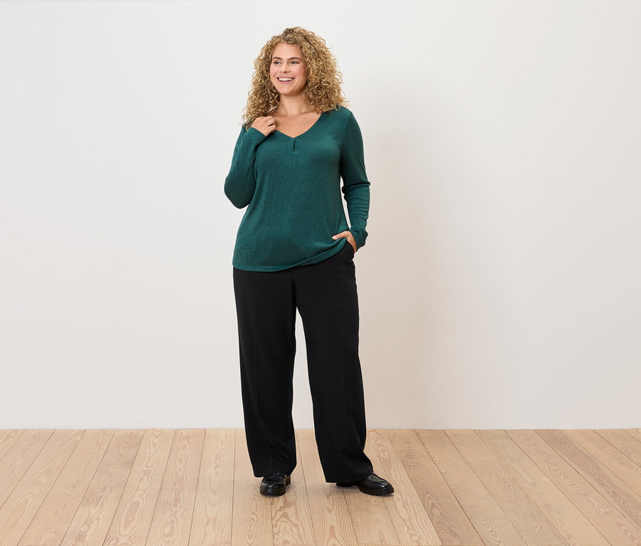 Žena s kudrnatými vlasy pózuje ve smaragdově zeleném svetru a černých kalhotách.