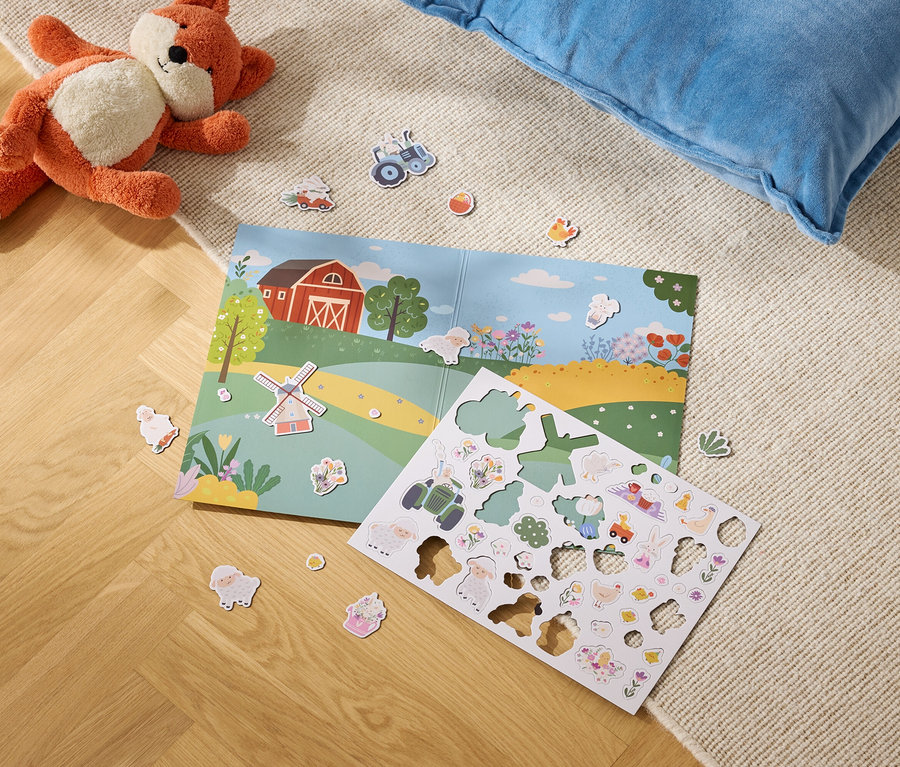 Plyšová liška leží vedle otevřené knihy s ilustrací farmy a archu s nálepkami farmy na koberci.