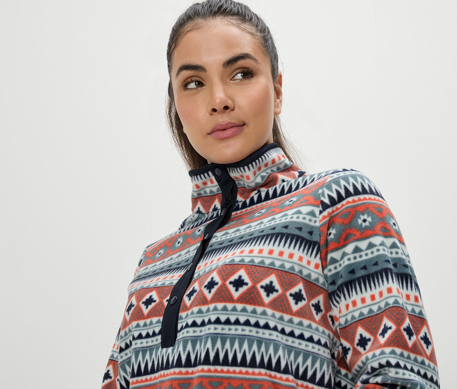 Portrét ženy s hnědými vlasy v pestrobarevné fleecové mikině s geometrickým vzorem.