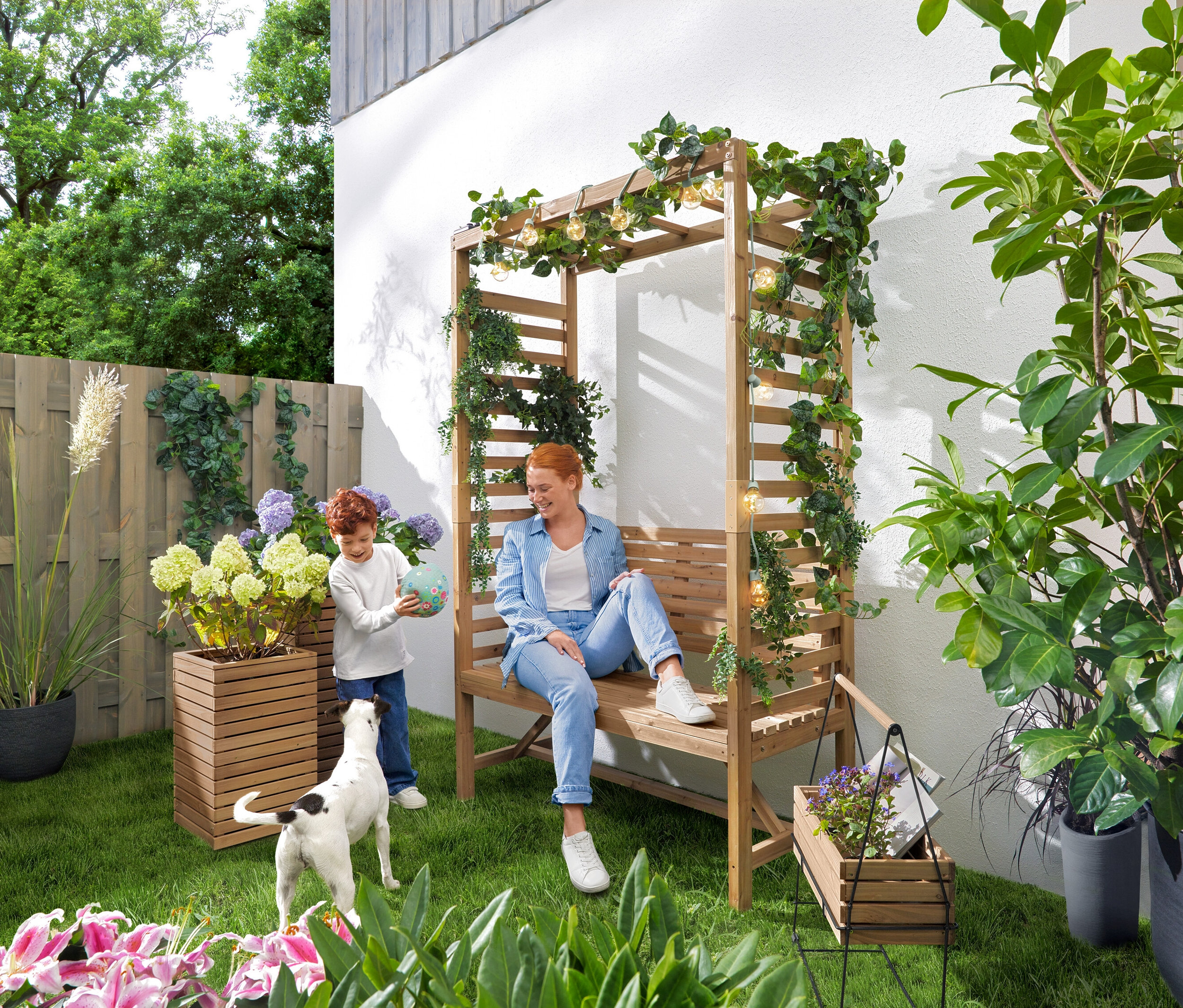 Žena sedí na zahradní lavičce s obloukem na růže, zatímco si dítě hraje s míčem. Nad lavičkou visí solární světelný řetěz »Žárovky«. V zahradě stojí truhlíky »Tinus«, 2 ks a truhlík na bylinky se stojanem »Elin«.