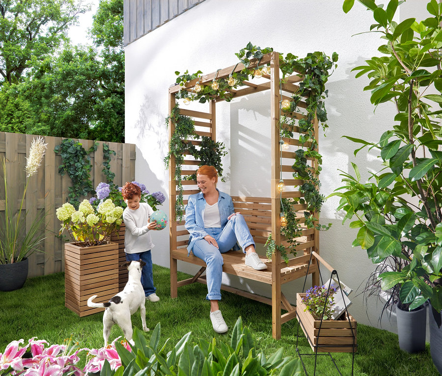 Žena sedí na zahradní lavičce s obloukem na růže, zatímco si dítě hraje s míčem. Nad lavičkou visí solární světelný řetěz »Žárovky«. V zahradě stojí truhlíky »Tinus«, 2 ks a truhlík na bylinky se stojanem »Elin«.