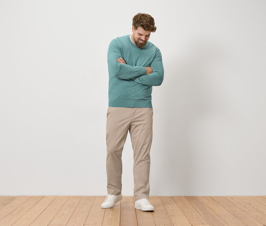 Muž s vousy stojí se založenýma rukama a má na sobě zelený svetr, béžové kalhoty a bílé tenisky.