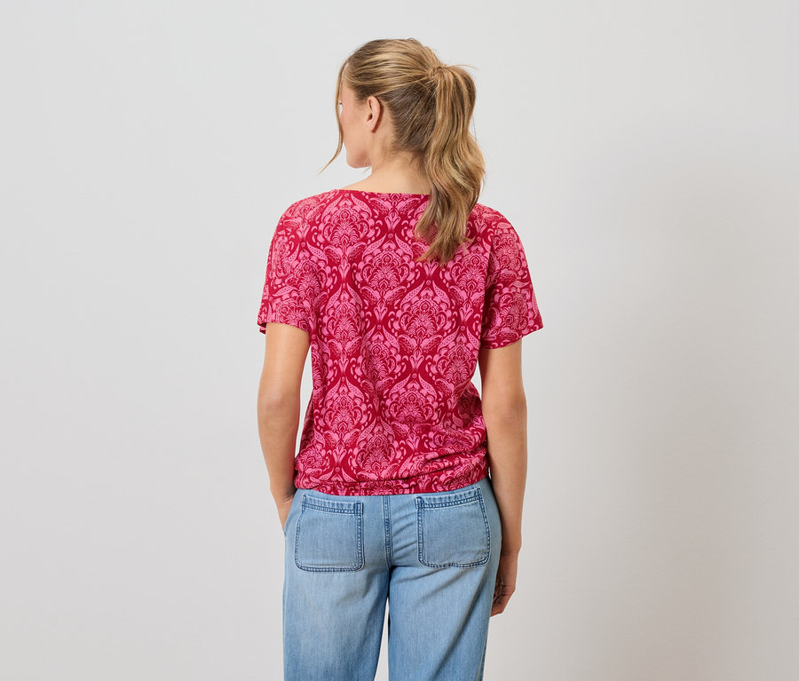 Žena s blond vlasy má na sobě červené vzorované tričko a modré džíny.