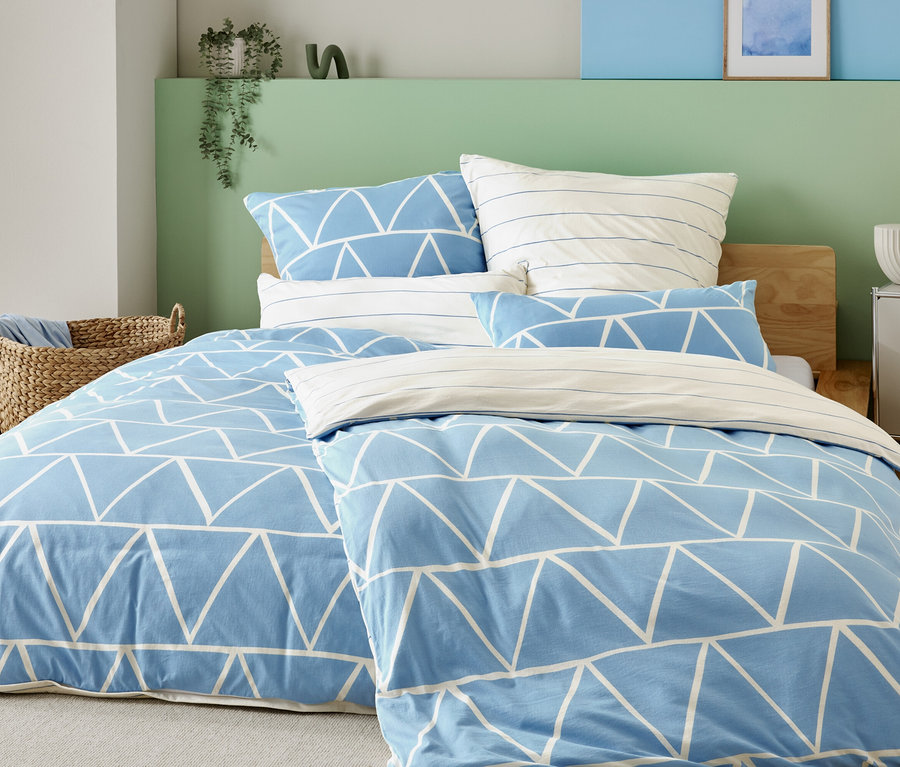 Postel se světle modrým potahem s bílými trojúhelníky a pruhovanými povlaky na polštáře, vedle proutěného koše a nočního stolku s lampou.