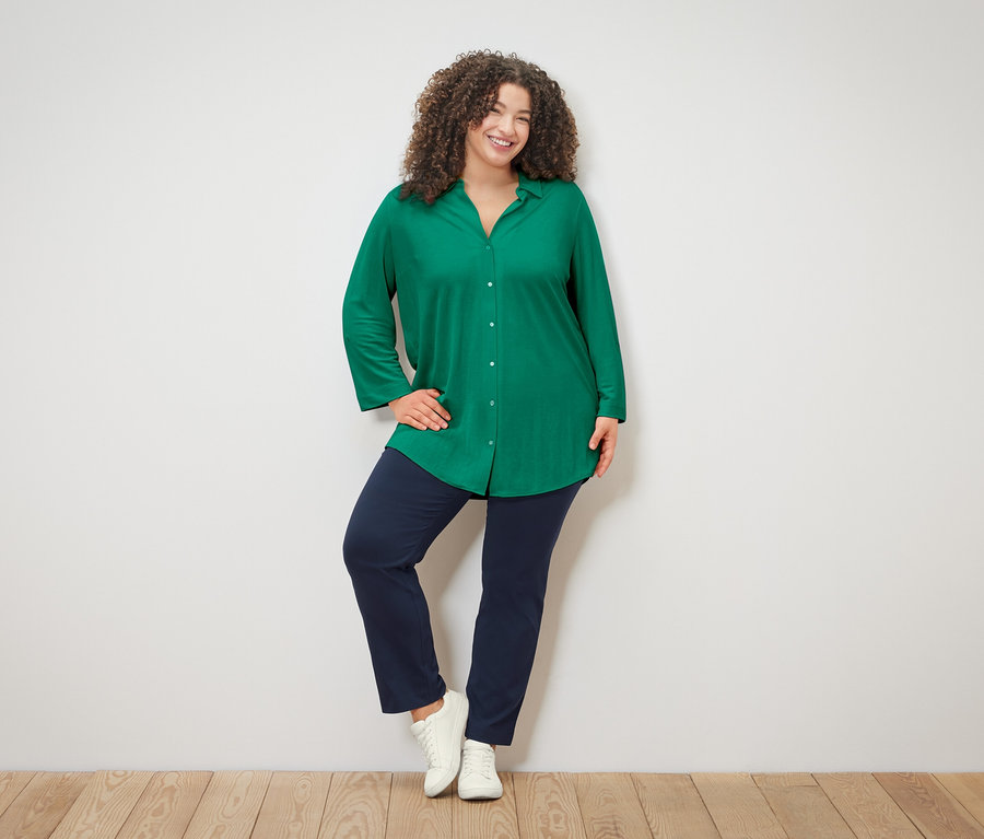 Žena s kudrnatými vlasy v zelené halence, modrých kalhotách a bílých teniskách pózuje na bílém pozadí.