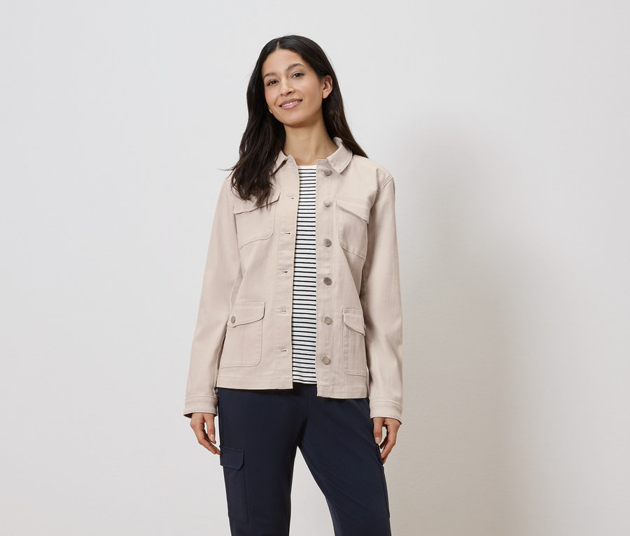 Žena má na sobě béžovou bundu přes proužkovanou košili a tmavě modré kalhoty.