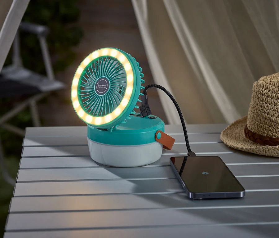 Tyrkysový kempingový ventilátor se světelným kruhem nabíjí smartphone na kempingovém stole, vedle něj je slaměný klobouk.