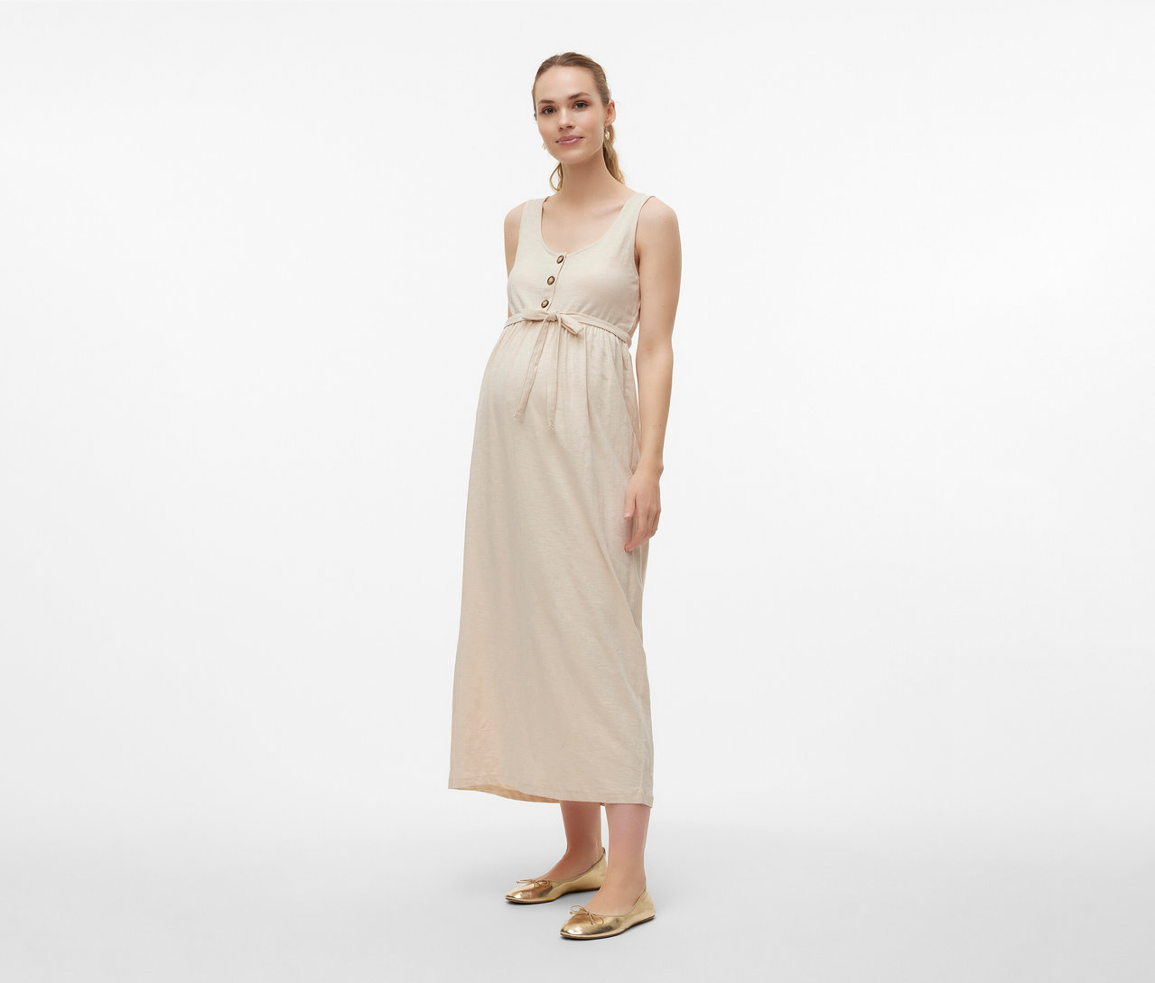 Žena nosí těhotenské šaty MAMALICIOUS »MLEVI LIA« s páskem a zlaté baleríny na světlém pozadí.