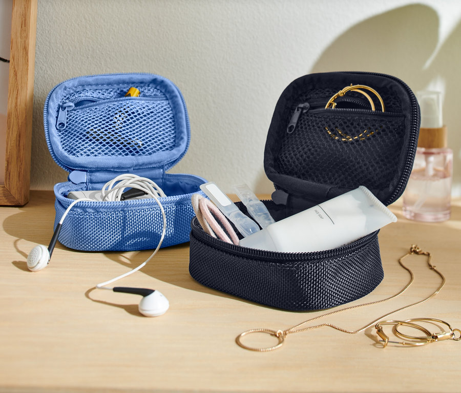 Na stole jsou dvě otevřené, malé úložné tašky, jedna modrá se sluchátky a jedna tmavá s produkty péče o pleť a šperky.