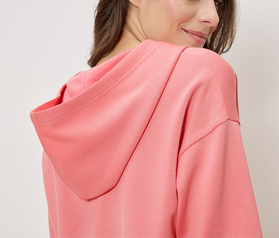 Žena má na sobě lososovou mikinu s kapucí, pohled z boku.