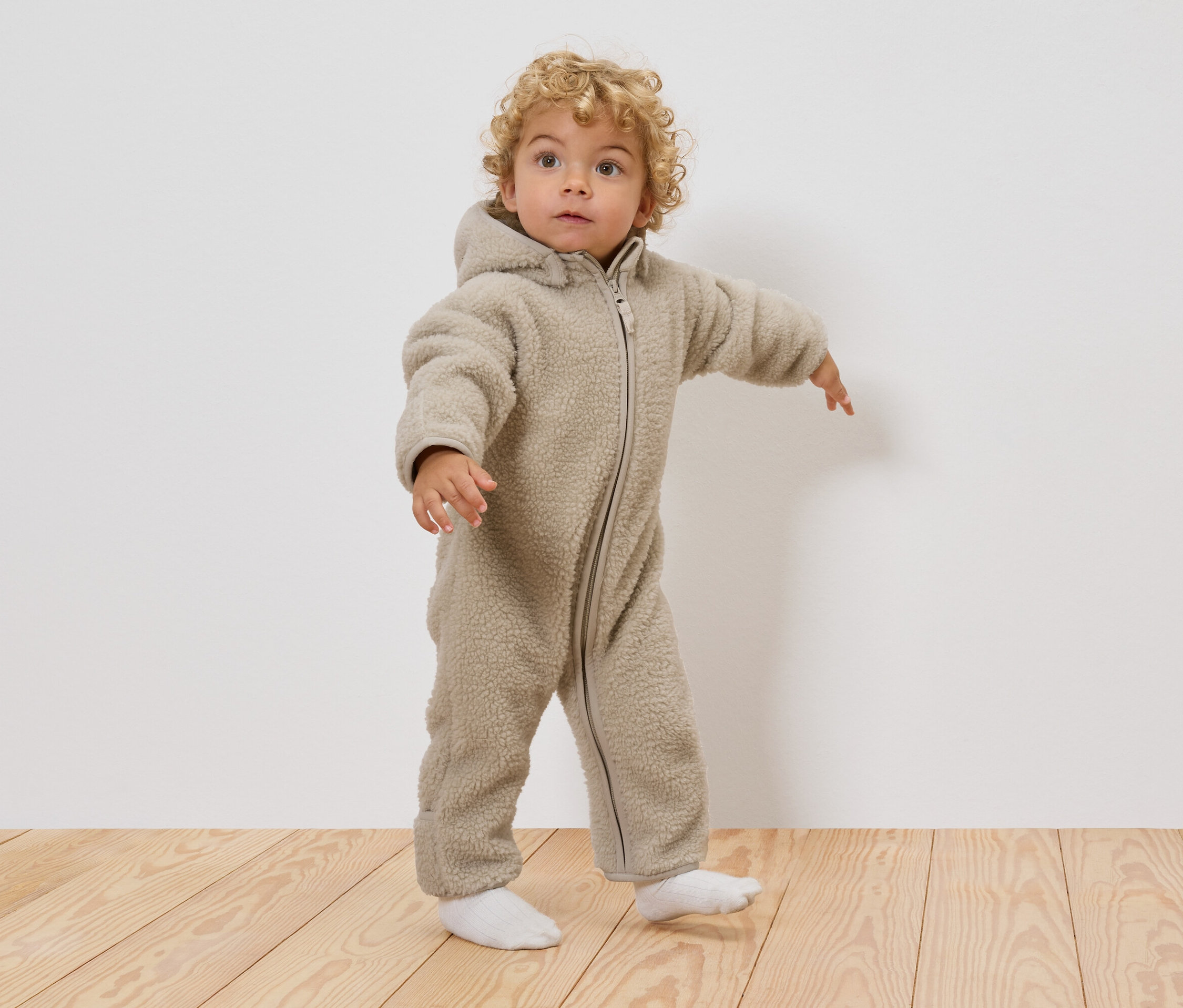 Malé dítě s kudrnatými blond vlasy má na sobě béžový teddy overal s kapucí a bílé ponožky, stojí na dřevěné podlaze.