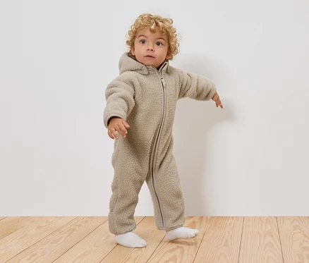 Malé dítě s kudrnatými blond vlasy má na sobě béžový teddy overal s kapucí a bílé ponožky, stojí na dřevěné podlaze.