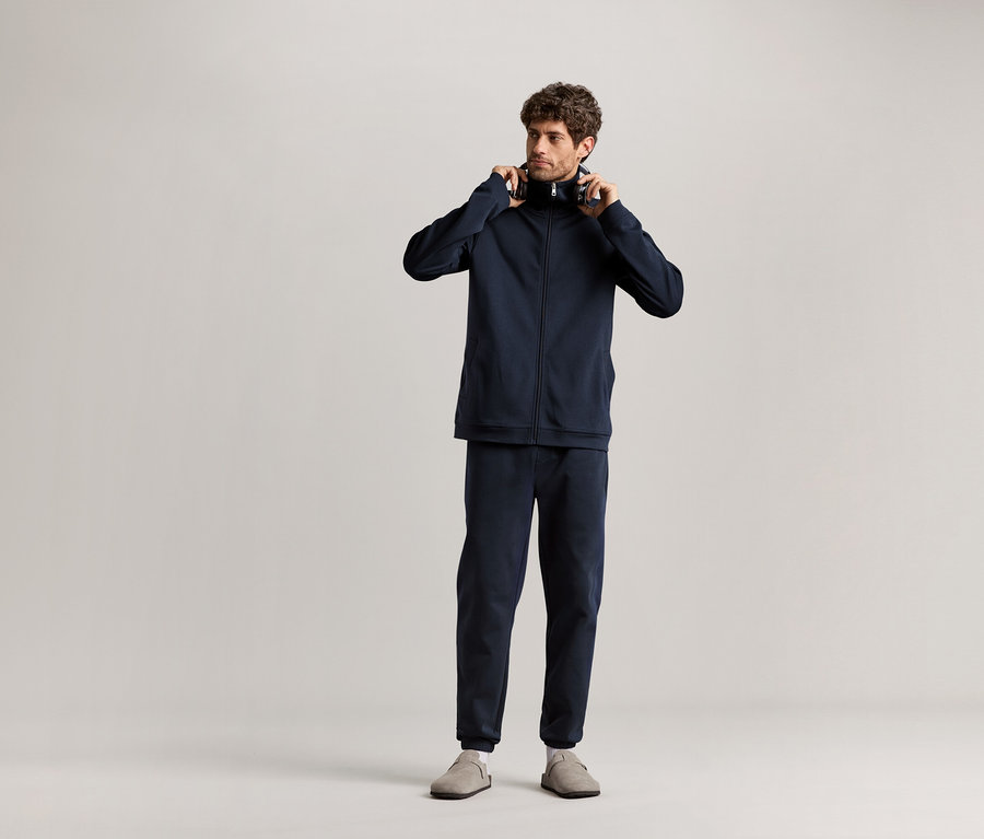 Muž stojí a drží sluchátka v ruce, má na sobě tmavě modrou domácí soupravu.