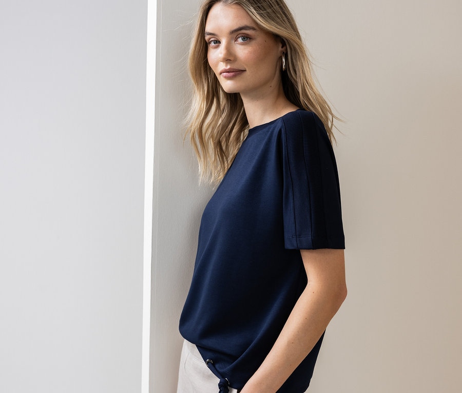 Žena se opírá o zeď a má na sobě tmavě modré tričko a světlé kalhoty.