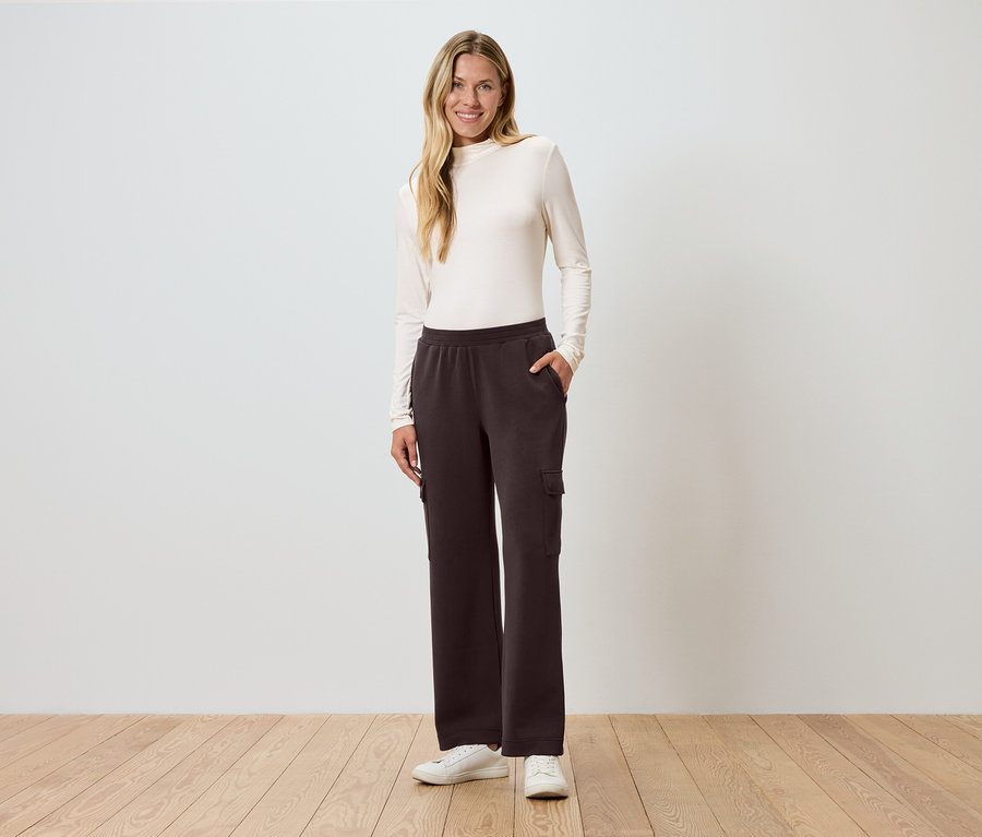 Žena stojí s rukou v kapse před zdí a má na sobě hnědé cargo kalhoty.