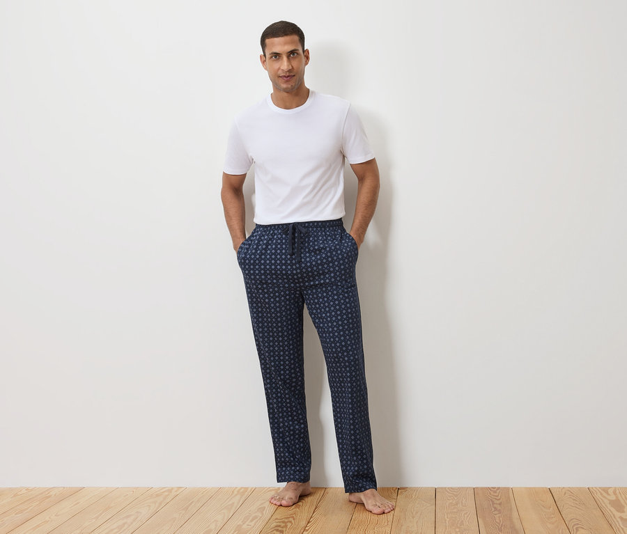 Muž stojí před bílou zdí, má na sobě bílé tričko a tmavě modré pyžamové kalhoty s puntíkovým vzorem.
