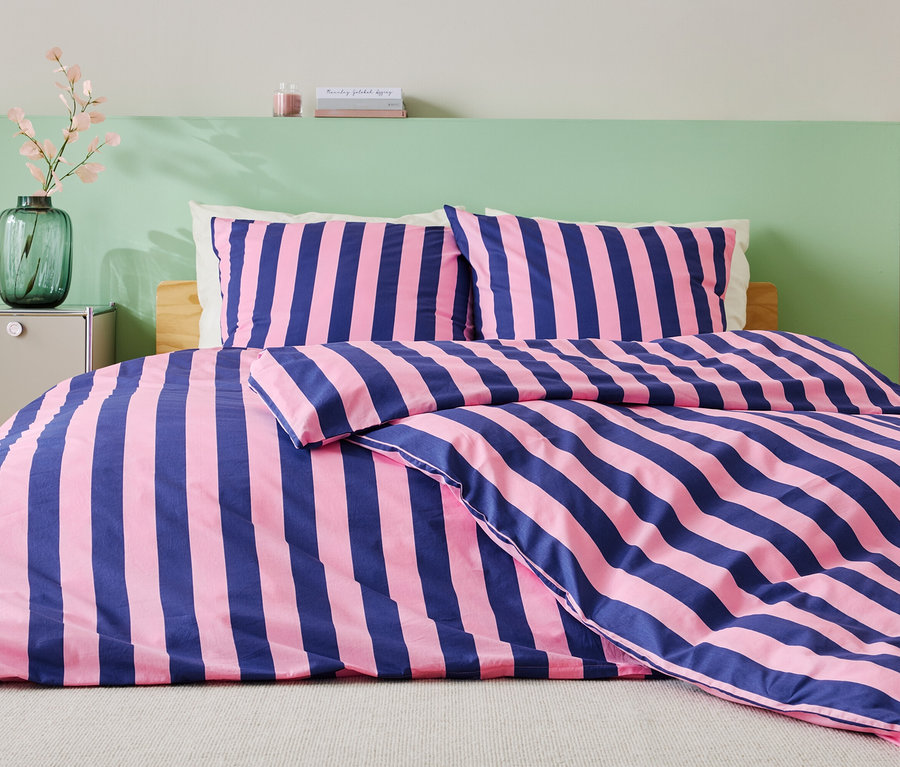 Postel s modro-růžovým pruhovaným ložním prádlem, dvěma polštáři a zelenou stěnou v pozadí.