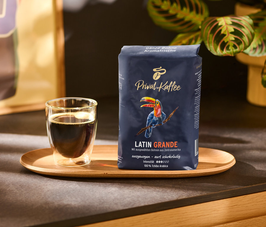 Šálek kávy a balíček zrnkové kávy Privat Kaffee Latin Grande na dřevěném podnosu.