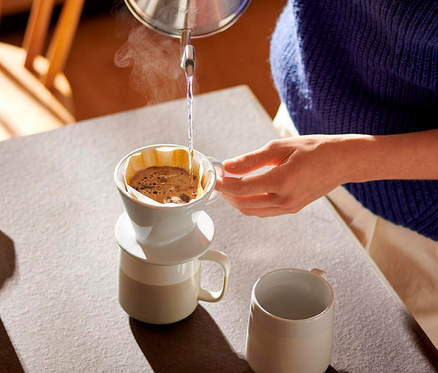 Osoba nalévá horkou vodu z konvice do bílého ručního kávového filtru, velikost 101, umístěného na hrnku. Z filtru stoupá pára. Na stole je další hrnek.