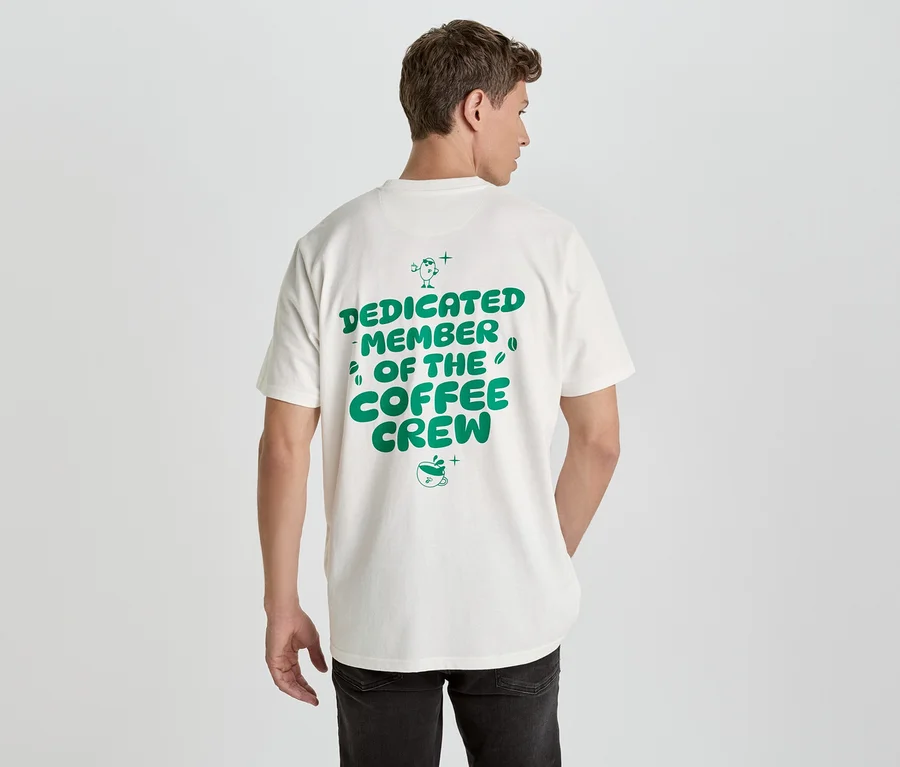 Muž zezadu má na sobě bílé tričko se zeleným nápisem 'Dedicated Member of the Coffee Crew'.