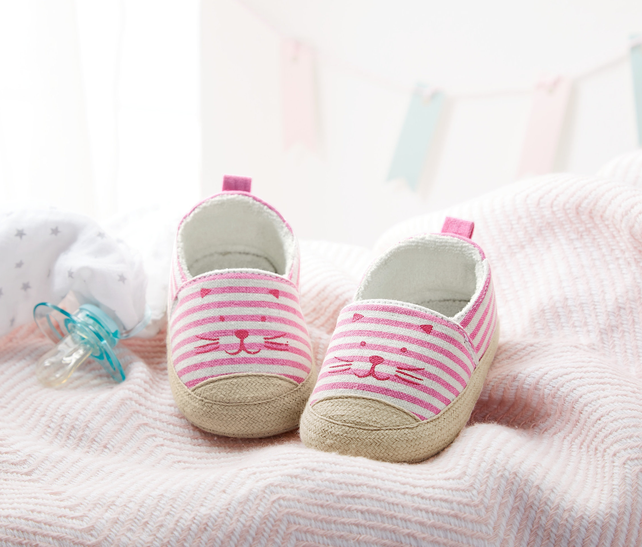 Dva dětské boty pro batolata s růžovými proužky a kreslenou tváří leží na růžové látce vedle modrého dudlíku.