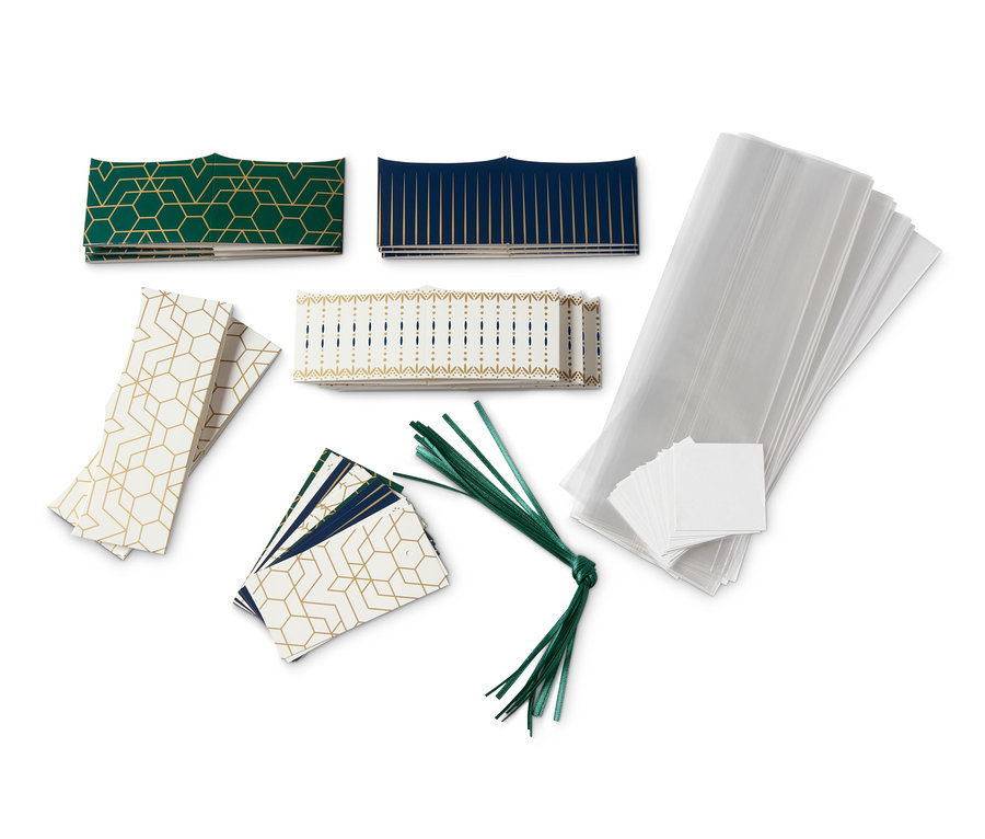 Obaly na cukroví s geometrickými vzory v zelené, modré a zlaté barvě, skládané na bílém pozadí.