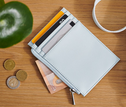 Na dřevěném stole leží světle modrá peněženka, mince a taška.