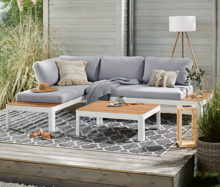 Terasa s šedou rohovou sedačkou, dřevěnou solární lucernou, světle šedým polyratanovým květináčem s nožkami a akumulátorovou stojací lampou s LED v dřevěném vzhledu.