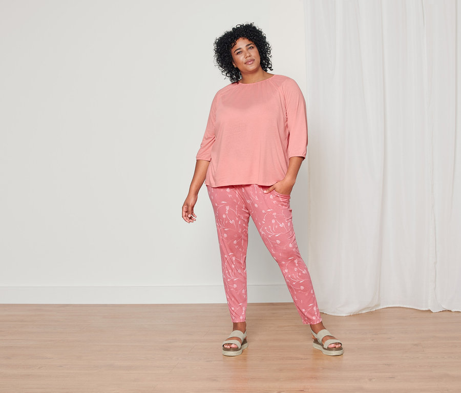 Žena pózuje v korálovém tričku s tříčtvrtečním rukávem, korálových žerzejových kalhotách s celopotiskem a kožených sandálech na platformě.