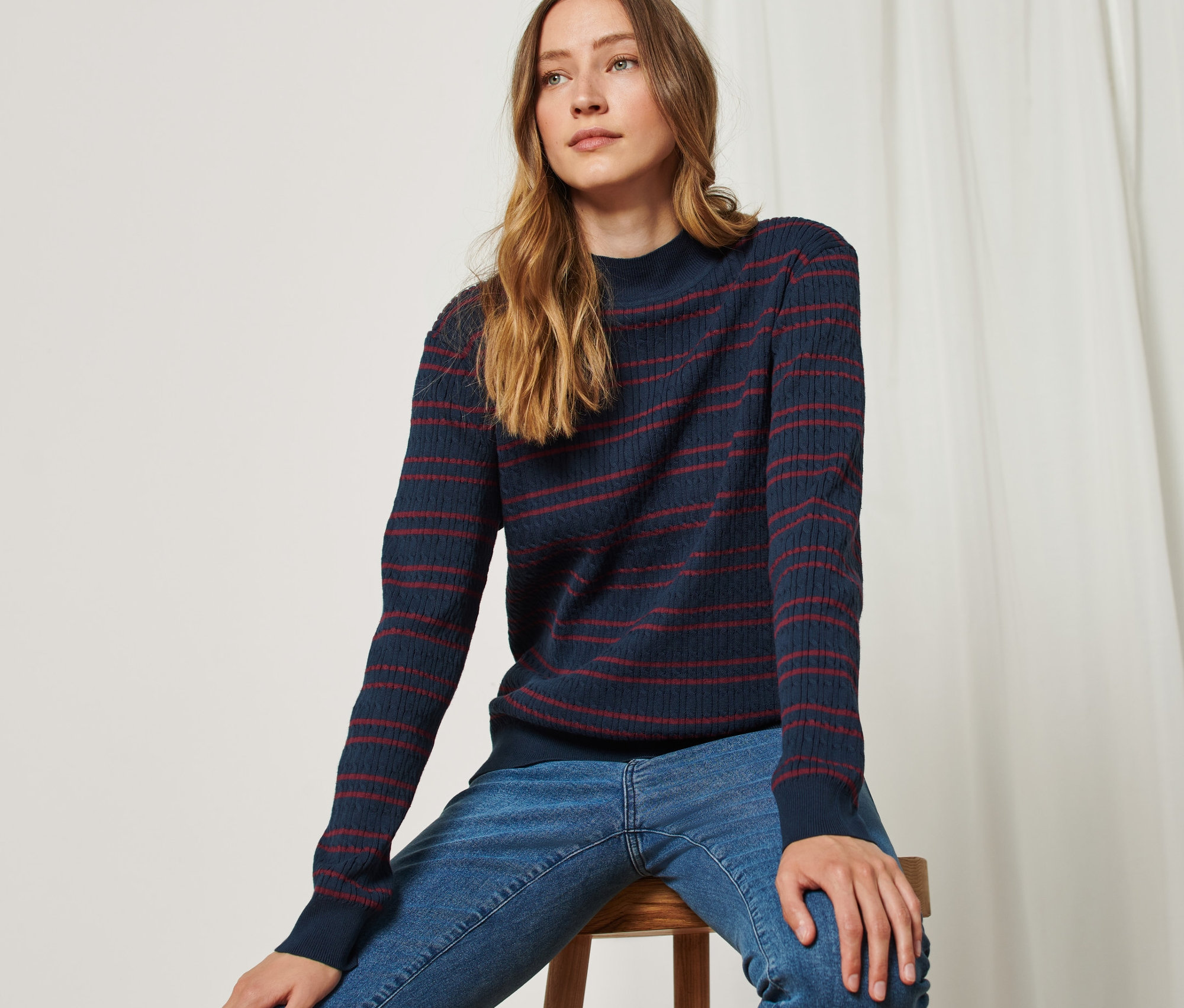 Žena sedí na stoličce a má na sobě modro-červený svetr z jemného úpletu s copánkovým vzorem.