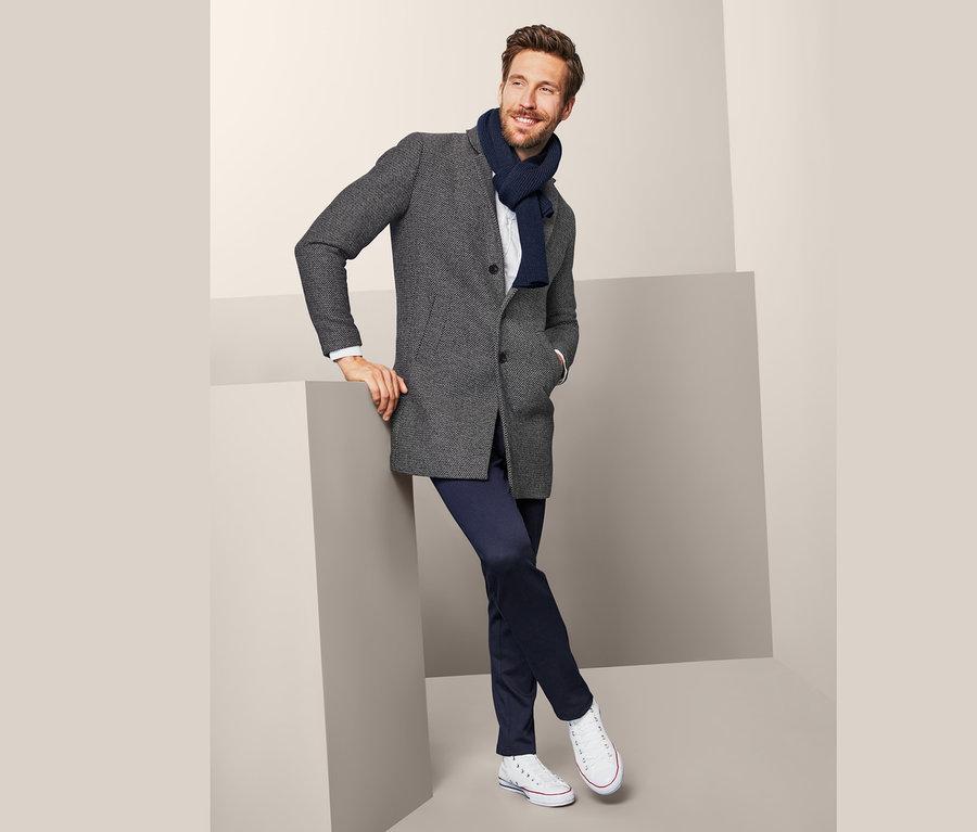 Muž opřený o šedou krychli má na sobě krátký kabát, modrou šálu, bílou košili, tmavě modré kalhoty a bílé tenisky.