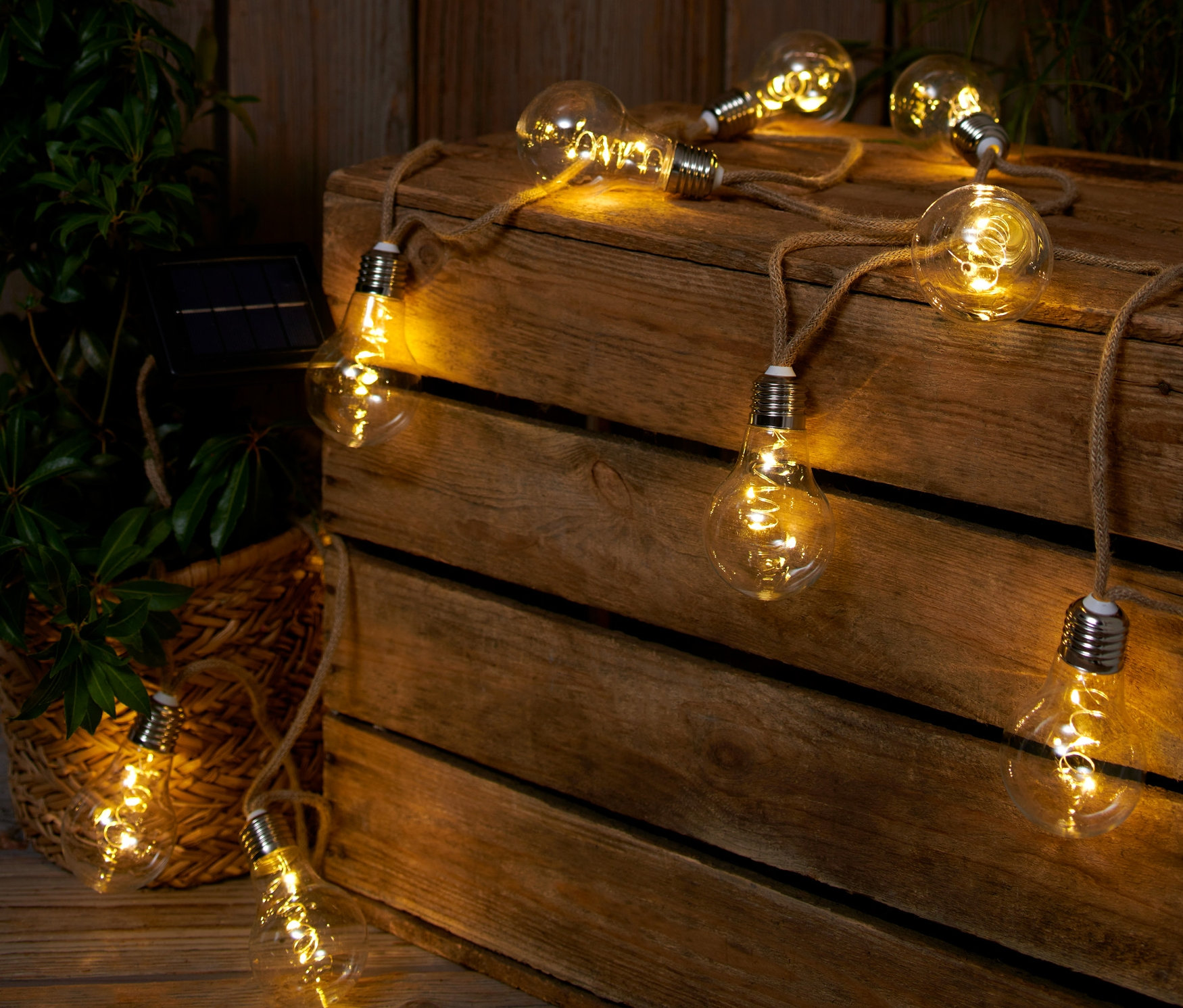 Solární světelný řetěz s jutovým provazem leží na dřevěné bedně vedle rostliny.