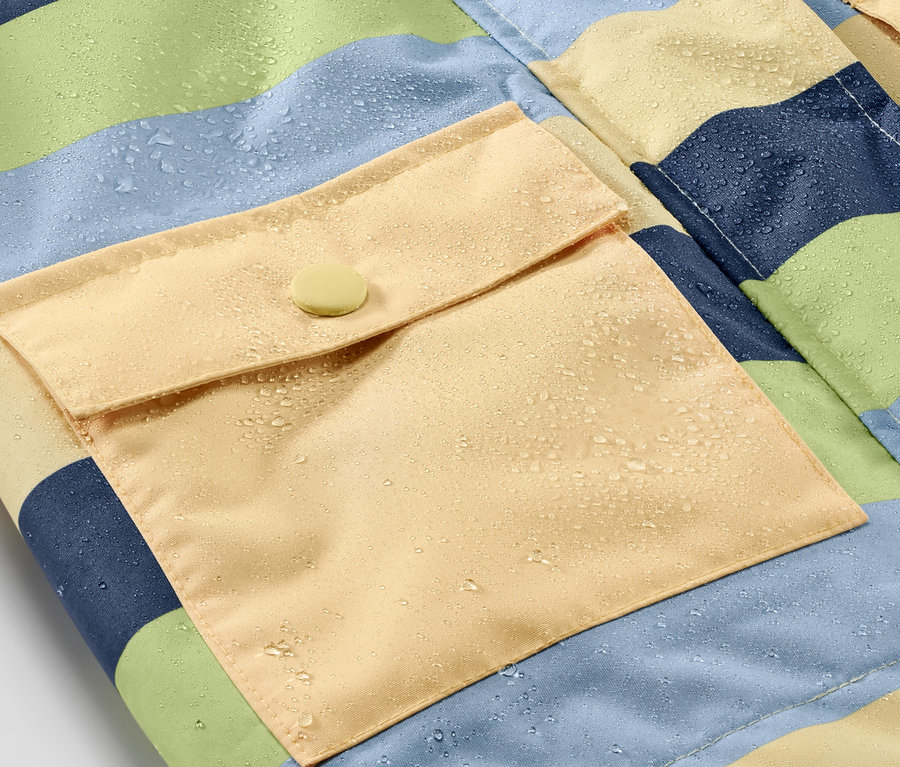 Detail dětské bundy do deště s proužky a žlutou kapsou s knoflíkem, na které jsou kapky vody.