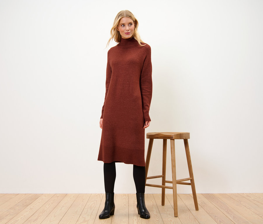 Žena stojí vedle dřevěné stoličky a má na sobě pletené šaty s vlnou.