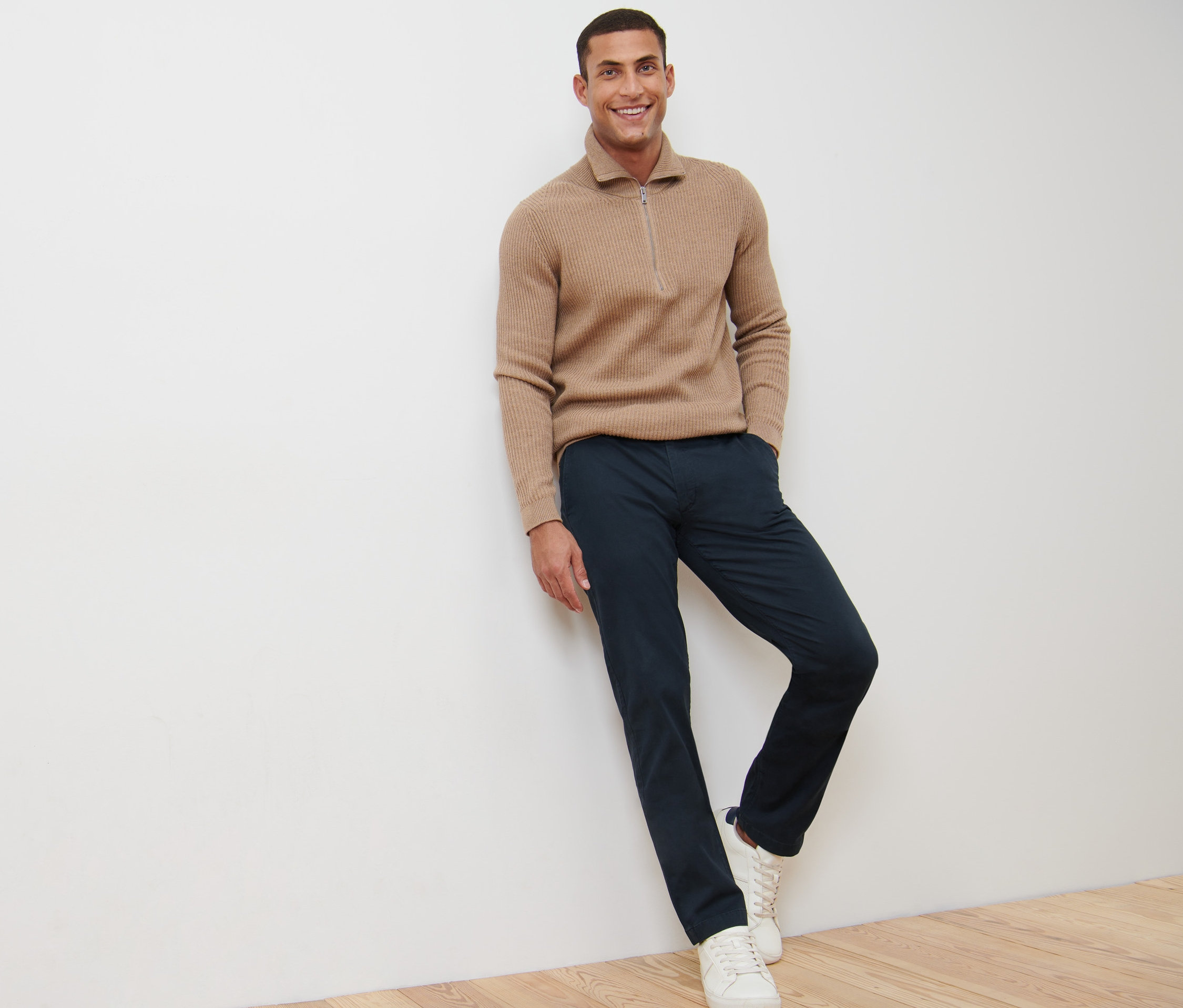 Muž se usmívá a opírá se o zeď v námořnických modrých kalhotách chino.