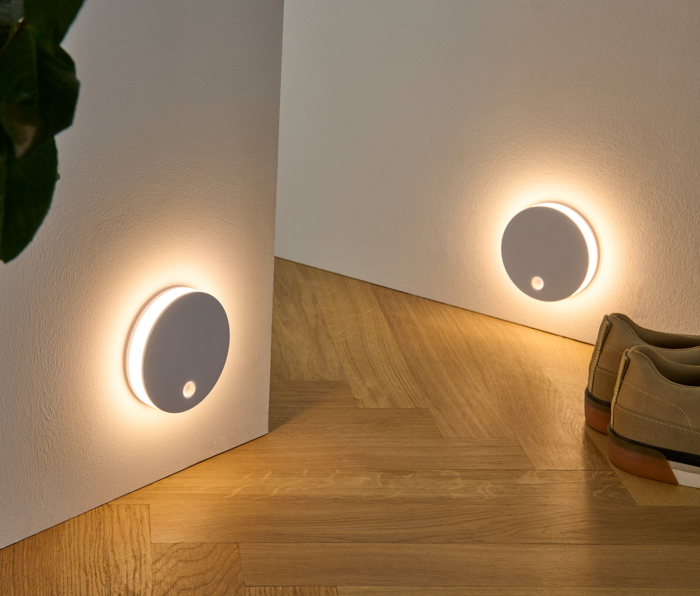 Dvě multifunkční LED světla připevněná na zdi, spolu s rostlinou v proutěném košíku a botami na dřevěné podlaze.