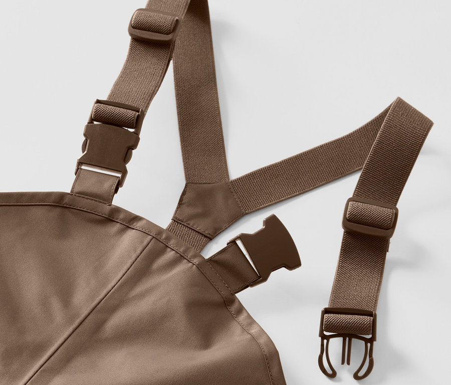 Detail dětských kalhot do deště s hnědými elastickými popruhy a plastovými sponami na bílém pozadí.