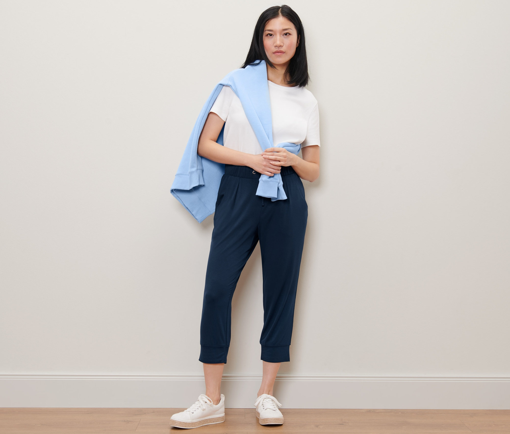 Žena stojí a má na sobě 3/4 žerzejové kalhoty, bílé tričko a přes ramena přehozený modrý svetr.