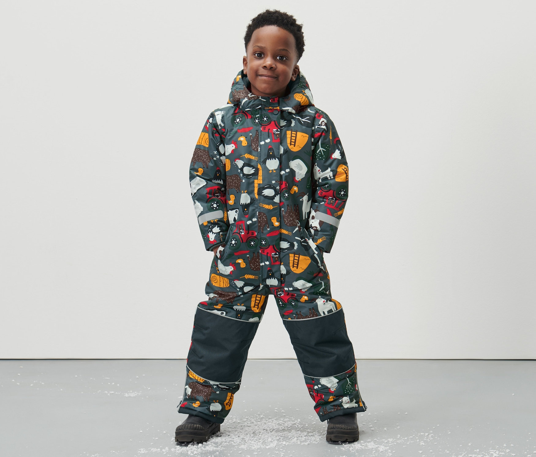 Chlapec v dětském zimním overalu s celoplošným potiskem farmy stojí na podlaze.