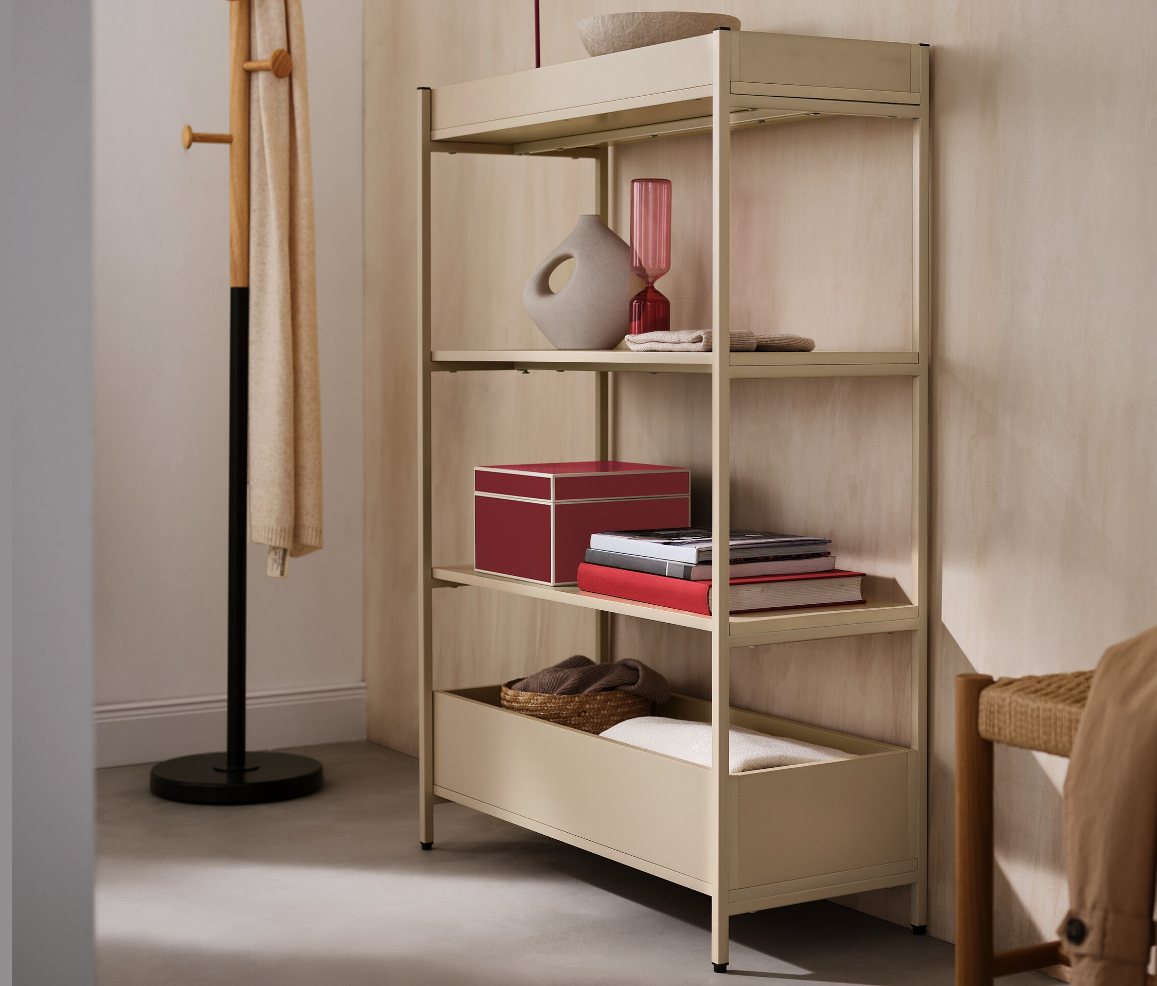 Béžová konzolová skříňka se čtyřmi policemi, na níž je červená LED stolní lampa, váza a miska. Vedle stojí věšák na šaty.