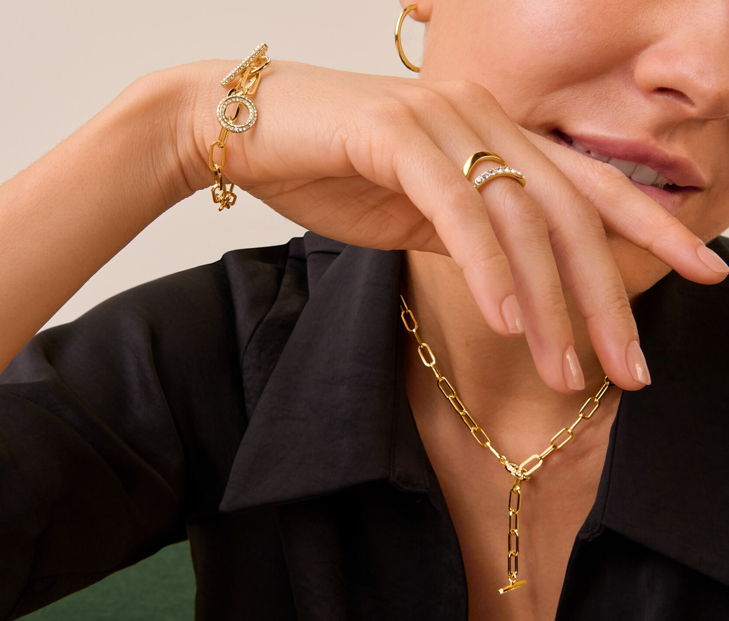 Žena se zlatým prstenem s dvojitou vlnou a kamínky, náhrdelníkem a náramkem Toggle osázeným kamínky a zlatými kruhovými náušnicemi.